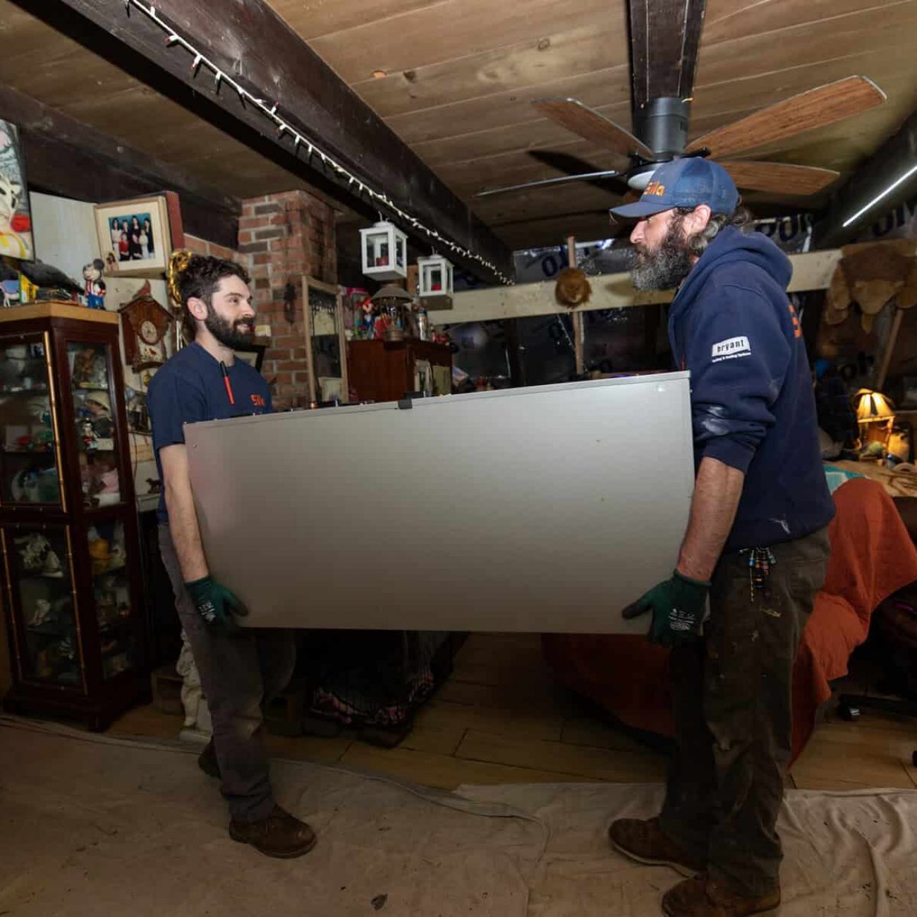 Two men wearing work gloves are carrying a large flat-screen TV through a cluttered living room with various decorations and furniture.