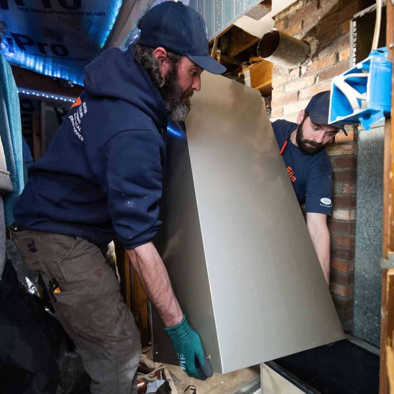 Two men install a large metal panel inside a partially constructed wall, surrounded by tools and construction materials.