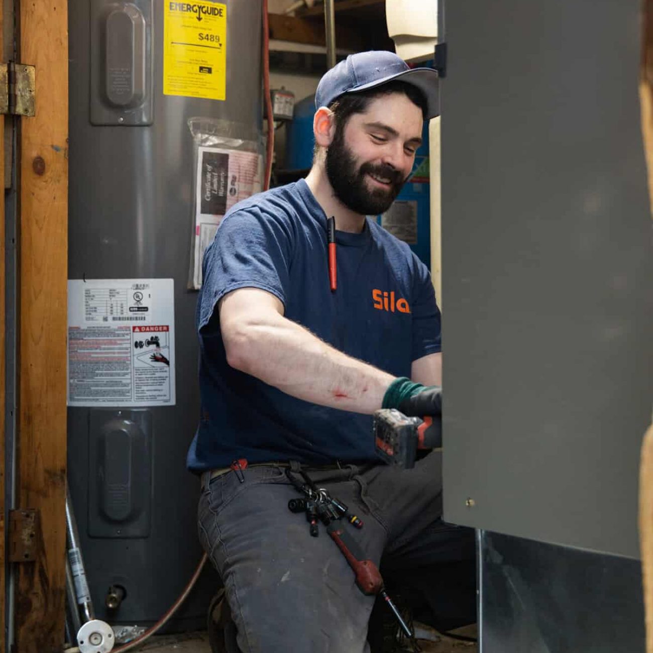 A technician in a blue uniform kneels while repairing equipment in a utility room with tools and a water heater visible.