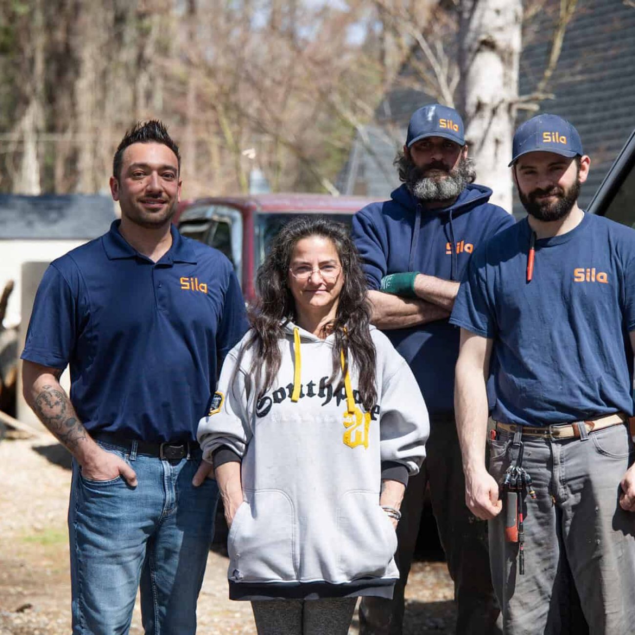 Four adults stand outdoors near a van and house, three wearing "Sila" uniforms and one in a gray sweatshirt, posing for a group photo on a sunny day during the Power the Nation Giveaway.